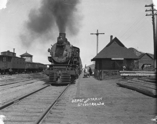 View down tracks towards a locomotive pulling a passenger train into the depot. A conductor stands in an open door. Another conductor and passengers stand on the platform. There are freight cars standing on tracks on the left in front of a water tower. Logs are piled in the foreground on the right. Behind the logs are two young children, one wearing a straw hat, the other a stocking cap. In the far background is a steep bluff.