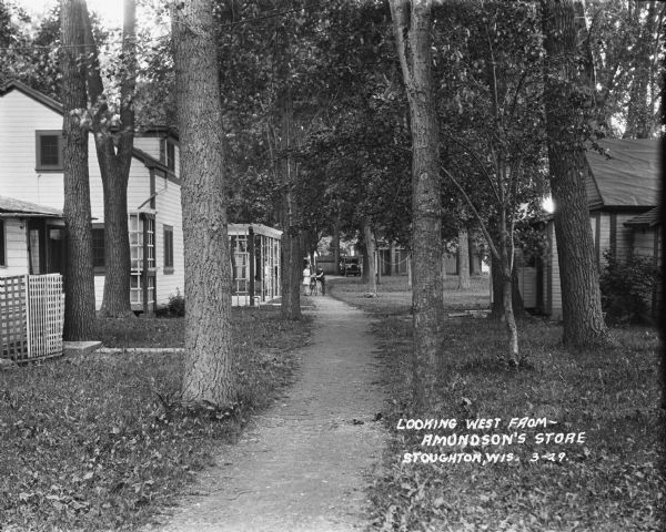 A view of small wooden houses on each side of a tree-lined path. Two children with a bicycle are standing on the path in the middle distance, near another boy standing under a trellis on the left.