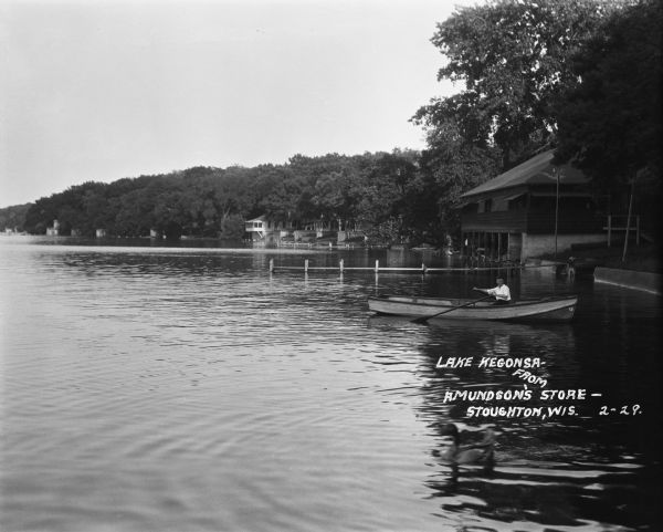 A view across water of Lake Kegonsa's shoreline with cottages, docks and trees. A boy is in a rowboat near a boathouse in the foreground.
