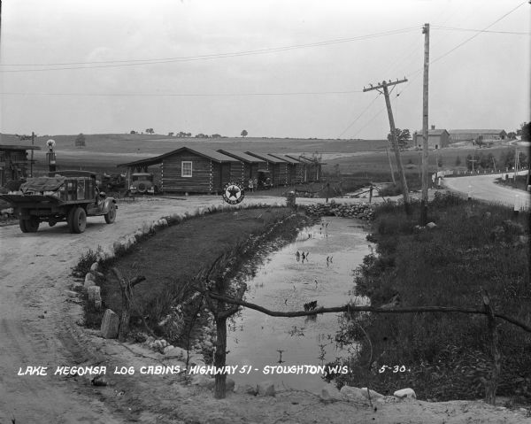 View from driveway over a stream towards a row of six log cabins to the left Highway 51. A Texaco Station and a delivery truck are on the far left. Cars are parked near the cabins. There are two girls near the second cabin. One is holding a pet. On the far right are farm buildings in the distance.