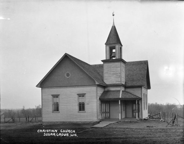 View of front of a wooden rural church, with a columned porch, belfry and steeple. There is a weather vane on the steeple. A wood and wire fence surrounds part of the church. In the background are fields and trees.