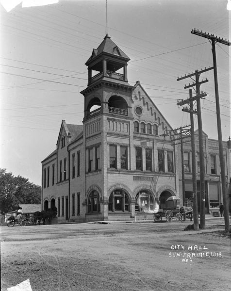 View across street of the Sun Prairie City Hall, a large brick building with two balconies in the tower and arched windows on the ground floor. A person driving a horse and wagon is parked on the left side. A horse-drawn wagon in front has an umbrella advertising for paint, and the horse wears a blanket that is advertising the State Fair. There is a drinking fountain on the street corner. A man is walking by a "seed buckwheat" sign posted in a store window.