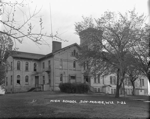 View across lawn towards the front of the high school, a large brick building with steps leading up to the main entrance, which has a tower above it. Two people are standing near the door. There is a fire escape on the left.