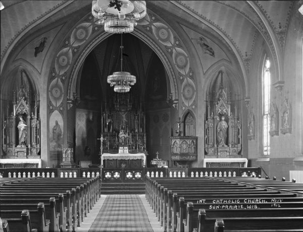 View down aisle between pews towards the altar in the interior of the Catholic Church. Ornate chandeliers hang from the ceiling. On the left in an arched niche is a Madonna and Child statue. On the right is an ornately carved wooden pulpit, and the other niche displays a statue of a saint, near a stained glass window. Angels are painted on the ceiling on either side of the altar.