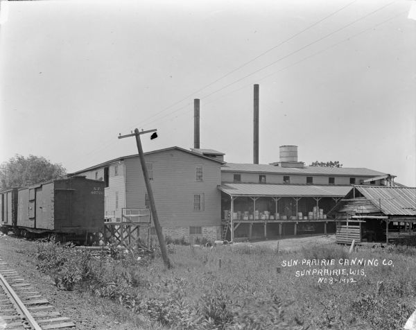 View from railroad tracks towards the back of the canning company. There are freight cars parked near the loading dock. Two smokestacks are behind the buildings. On another loading dock, which has a roof, a man is standing near stacked metal tubs.