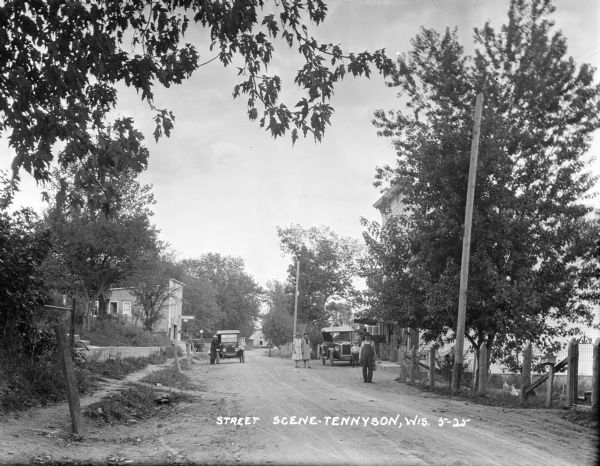 View down street towards a garage with a gas pump on the left. There is a man standing by a parked car. Two women and a boy are standing near a parked car on the right. A man with a jug is in the foreground standing next to a fenced-in yard with chickens.