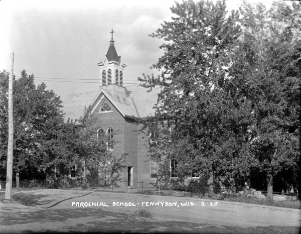 View across road towards the parochial school, which is a two-story brick building with a cross on the steeple. Arched windows are above the entrance. Trees are on both sides of the entrance. There is fencing around the property. A girl stands under a tree on the right.