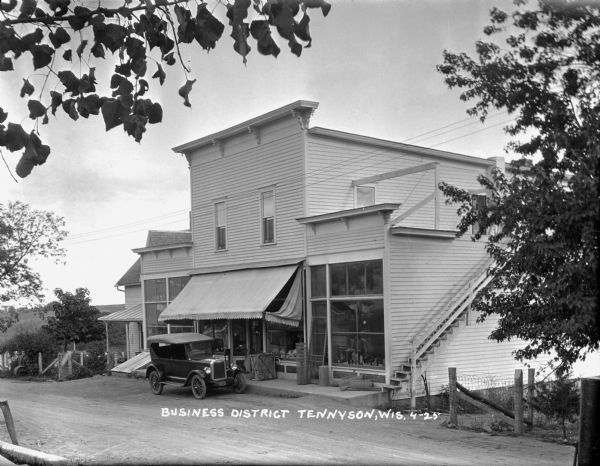 View across street of the business district towards a general store, which has merchandise displayed in the large front windows. An automobile is parked in front. A flight of stairs on the right side of the building leads up the to the second floor.