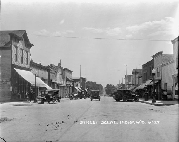 A view of downtown Thorp. Lampposts lining the street on both sides. Cars parked at the curbs on both sides. A couple cars driving away down the street. On the left there's a general store, a bank and a hotel. On the right there's a gas pump and Hotel Thorp. Some pedestrians on both sides.