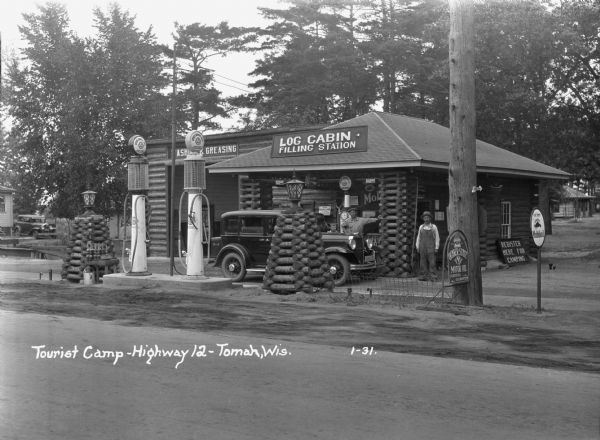 A view of the Log Cabin Filling Station at a tourist camp. A garage for washing and greasing on the left. Two tall gas pumps. Lamps made of interlaced logs. A man and boy standing behind a car. An attendant standing at the door. Several signs for oils and gas products.