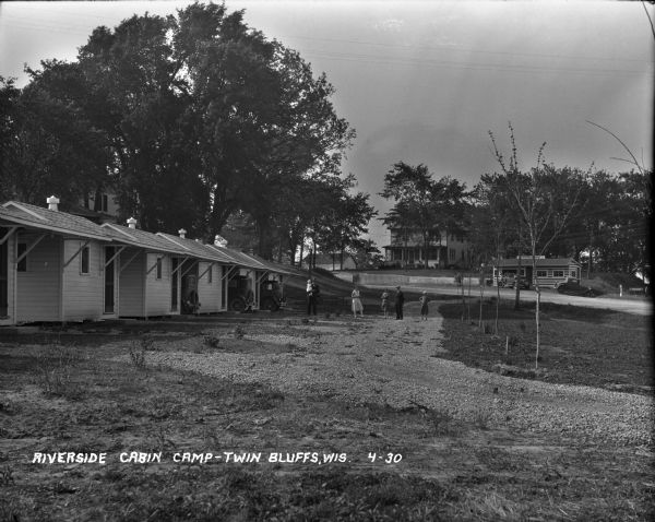 A view of a camp with a row of small cabins on the left. Automobiles parked between the last three. Some people standing in the driveway -- a man with a baby, two women, another man and a boy. A building with a gas pump across the road with two cars parked in front. A house with a large porch behind that.