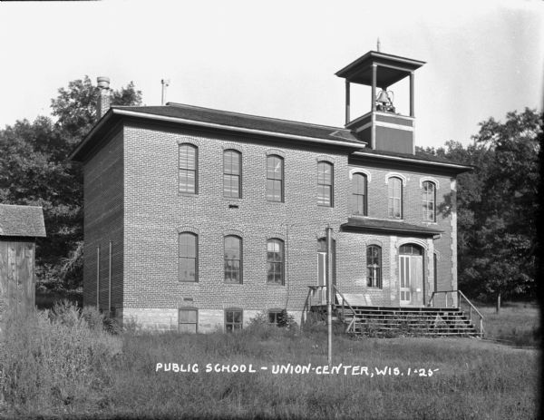 The public school, a two-story brick building with tall windows in the front but not on the side. A bell above the entrance. A shed to the left.