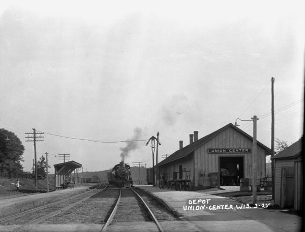 View down tracks towards a depot with an oncoming train arriving from the opposite direction. On the left side of the tracks opposite the depot is a shelter with benches. A railroad worker and a row of hand carts are on the platform.