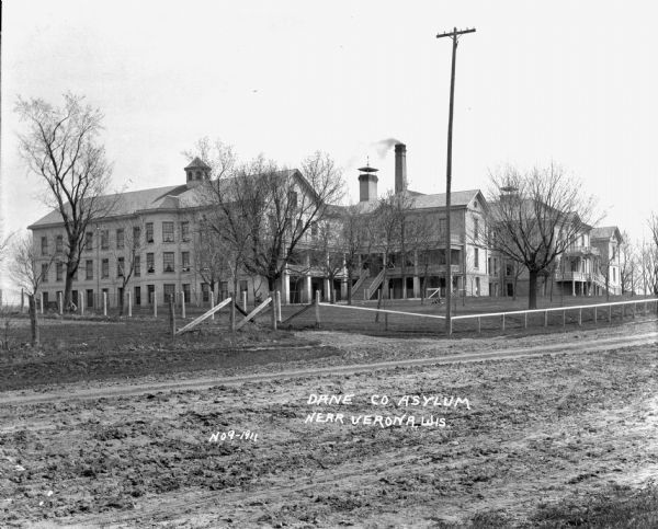 View across dirt road of the asylum and grounds. A large three-story building is spread out over several acres. Stairs are leading up to entrances on the second floor. Three people are on the lawn in front behind the fence. One man is lounging in the grass, another person is sitting on a bench, and the other person is sitting under a tree.