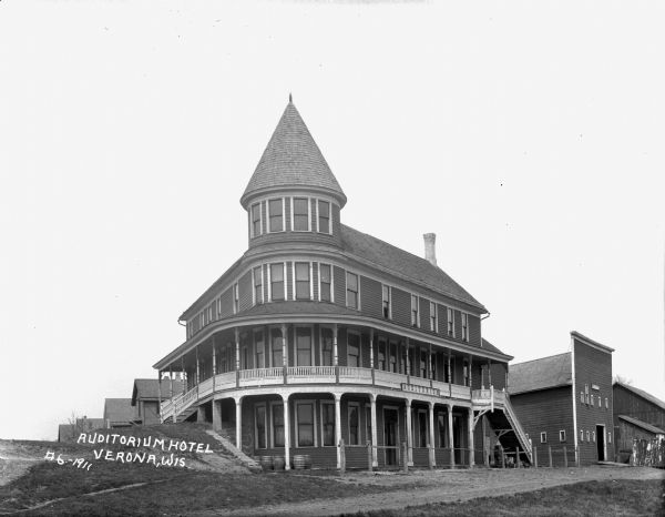 The Auditorium Hotel, a three-story building with a turret and stairs to the main floor entrances. A wrap-around balcony is on the main floor. Barrels under the windows on the ground floor, and a row of buildings are behind it on the left. There is a stable to the right.
