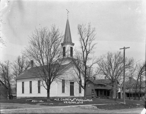 View across street of the M.E. Church, which is a one-story wood frame building with a weather vane on the steeple. There are tall windows on either side of the entrance and along the side. The parsonage is behind on the right. There is a cat on the porch. A house with a wrap-around porch is in the background.
