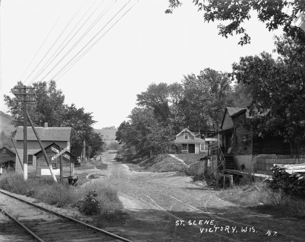 View down road over railroad tracks towards buildings and dwellings on the left. A family is standing on the stoop of a house in the far background on the left. The school with bell tower is at the end of the street. On the right is a post office with stairs leading down to a boardwalk and the street.