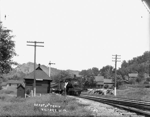 View over railroad tracks towards a railroad depot with an arriving train. A few passengers are on the platform. On the right is a pile of fuel wood. There is a bluff in the background.