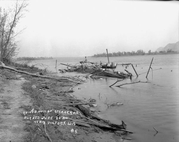 A man stands on the wreckage of a burned steamer in the Mississippi River near the shoreline. There is a small boat and a barrel nearby. Bluffs are in the distance on the right.