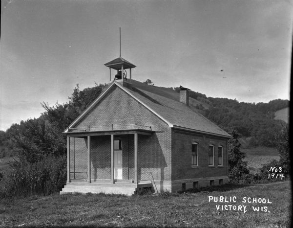 Exterior view of a small brick public school on a hillside. There is a bell in a small bell tower above the entrance.