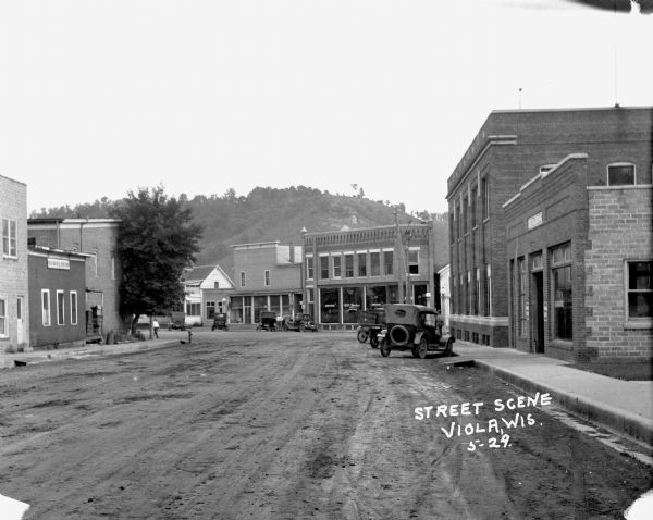 View down unpaved street scene towards an intersection. A tree and a fire hydrant on the corner on the left. On the right is a garage with a ramp entrance at the sidewalk. Trucks and cars are parked at the curbs. Across the intersection is a drugstore with large display windows. In the background is a steep bluff with trees.