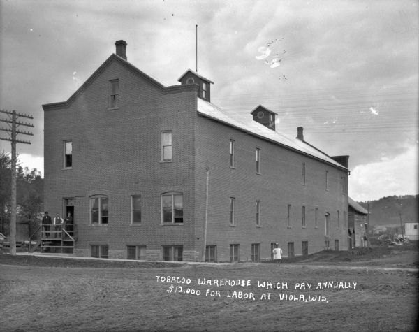 View across unpaved road of the front of a brick two-story tobacco warehouse. Three men are posed standing on the stoop in front of the door. A barefoot girl is standing on the sidewalk on the right. In the far background is a tree-lined hill.