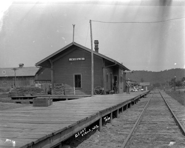 View down center of tracks along platform towards the railroad depot. Two men are standing on the platform near two carts. On the platform are two wooden crates which read on the side: "Potosi Brewing Co." A  large pile of what may be bricks are stacked behind the depot on the left near barns and industrial buildings. There is a tree-lined bluff in the far background.