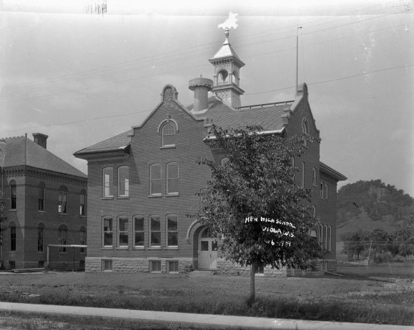 View across lawn of the high school, which is a two and a half story brick building, and on the roof is a large round metal vent, and a belfry with a copper roof. The double door entrance is in an archway. Trees on a bluff in the background. A bicycle is leaning against the two-story brick building next door.