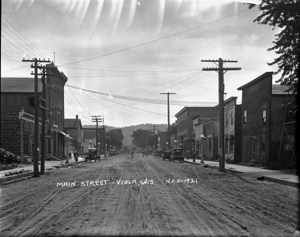 View down center of unpaved Main Street, with a service garage on the left and ice cream parlors on the right. There are lampposts along the sidewalks, and cars are parked along the curbs. On the far right is what appears to be the base of a metal tower, and in the far background is a tree-lined hill.