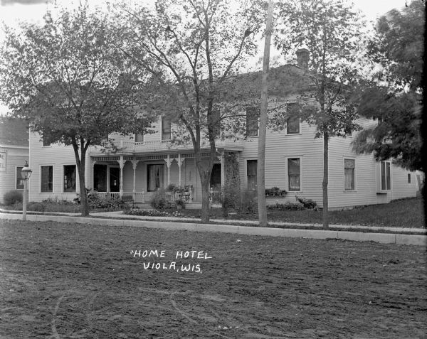 View from unpaved road of the "Home Hotel," a two-story wood frame building with a balcony and a porch. Several flowers are planted in the garden and in pots between the windows. There are young trees along the curb, and a lamppost in the street in front of the entrance.