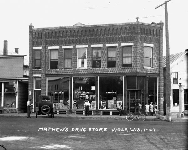 View from street of Mathew's Drug Store, which is a two-story brick building with large display windows on the first floor. A number of people are on sidewalk in front, and there is a woman standing at one of the second floor windows. There is a drinking fountain at the curb near a lamppost with a 56W sign. A bicycle is parked at the curb. On the left is a shop that sells hats and shoes.