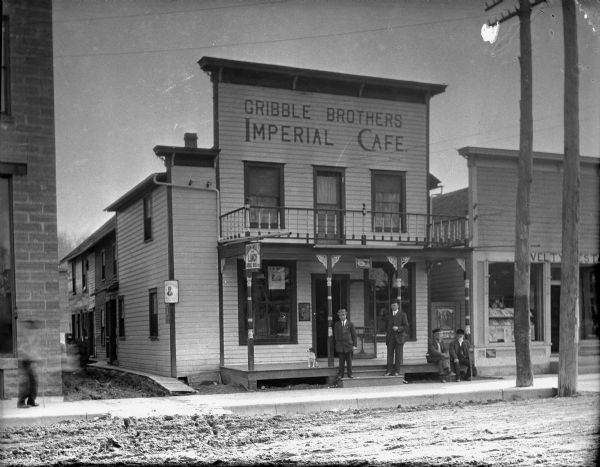 View from unpaved street of the Gribble Brothers Imperial Cafe, which is a two-story wood frame building with a small porch and balcony. Two men are standing on the front stoop, and a dog and two bearded men are sitting on the porch. There is an alley on the left, and a novelty store on the right.