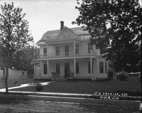 View from street of the J.H. Frazier Residence, which is a two-story wood frame home with a porch and a balcony. There is a small garden in the center of the front walkway. On the porch are two chairs and a settee. There is a hand-pump to the left of the porch.