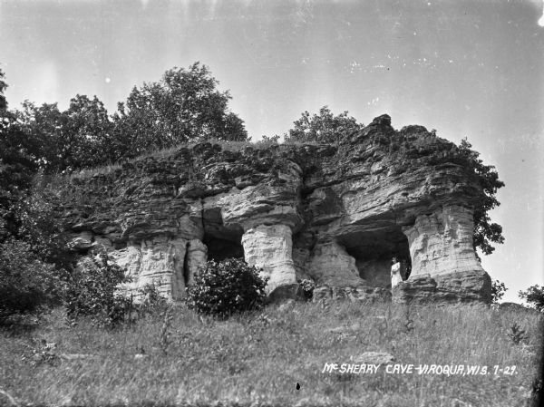 View uphill towards the entrance to the McSherry Cave. A woman is standing at the entrance. Trees and shrubs are growing on top of the cave.