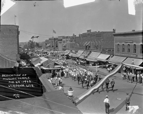 Rooftop view of a parade passing through downtown Viroqua. A boy is sitting on a roof on the left, looking down towards the street at the marching bands and Freemasons. Spectators are on both sides of the street, and a tent in the street is decorated with flags and banners, probably for concessions. Just above street level is a cut-out of Harold Lloyd's head on the building next to the drug store. A stationery shop, hardware store and other businesses are on the right.