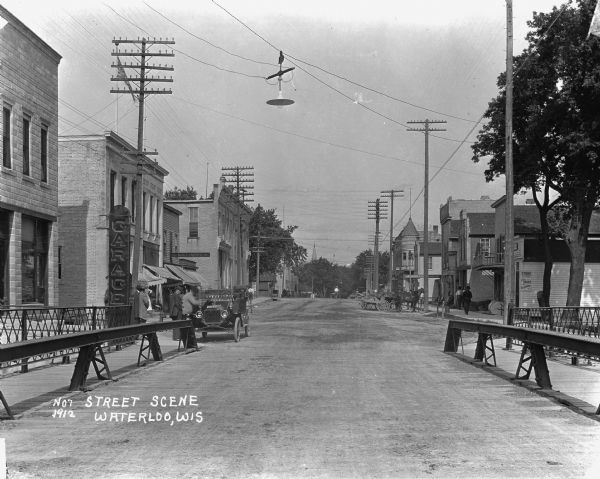 View down street from the end of a bridge. On the left is a service garage, post office and millinery. On the right is a shoe and repair shops, and other commercial buildings. An automobile is parked on the left with a group of men standing nearby. Horses and wagons are parked on both sides of the street.