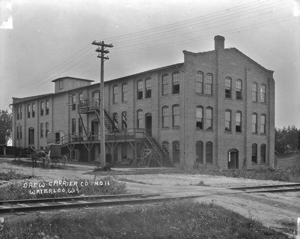 View across railroad tracks of the Drew Carrier Company, which is a three-story brick building with exterior stairs and a ramp leading up to a second floor loading dock. A horse and buggy are parked at the curb in front. A man is standing at the second floor corner window, and a dog or pig is standing in the grass near the electrical pole.
