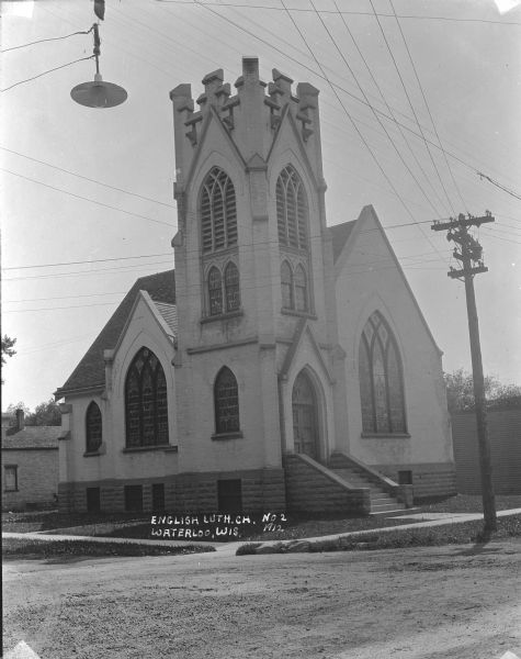 View from street of the English Lutheran Church on a corner, showing front and left side of the building. The entrance is under a square bell tower, which has no steeple. There are large, arched stained glass windows. A street light is suspended over the street on the left.
