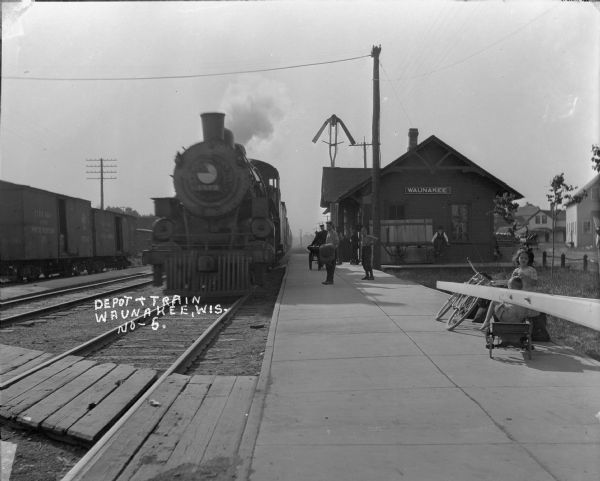 View down platform toward the Waunakee depot and an arriving train. Freight cars are on the tracks on the left. A group of men are standing on the platform, and a bearded man sits on a cart at the side of the depot. In the foreground on the right are a girl and boy with a coaster wagon and a bicycle, just behind the guard bar that is down to block the crossing.
