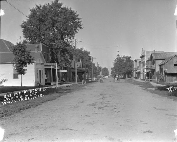 View down Main Street, with a railroad crossing at the far end of the street. On the left is a shoe repair store, lumber business, and a lunch room with a girl standing at the entrance. Unidentified businesses are on the right. There are a group of boys standing in the street. A horse and carriage are approaching up the street.