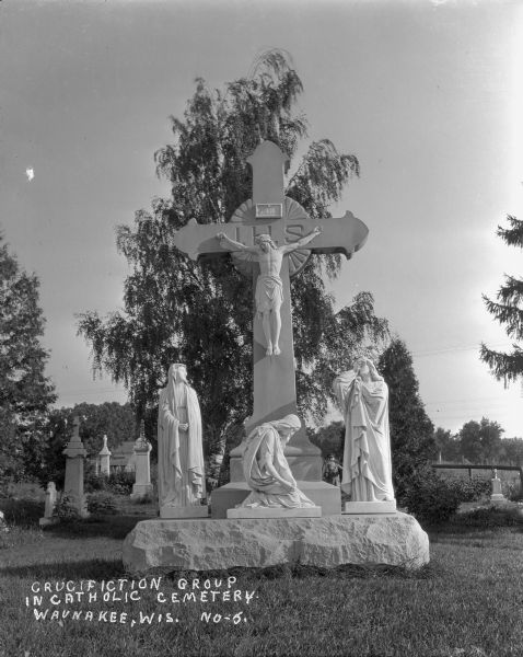 A marble crucifixion monument of Christ and three mourners in the Catholic Cemetery. A groundskeeper stands just behind the statue holding up a scythe. Other marble grave markers are visible in the background.