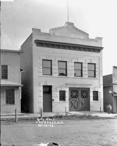 View from across street of the Village Hall/Fire Station. There are hitching posts along the curb. On the right a young boy stands on the sidewalk near a farm implement store.