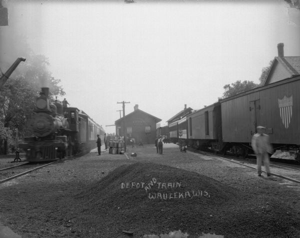 View towards railroad depot with trains on railroad tracks on the left and right. In the foreground is a pile of gravel. There is a group of people on and around the platform on the left, and there is a large stack of milk cans at the end of the platform. There is a railroad worker standing on the top of the locomotive on the left.