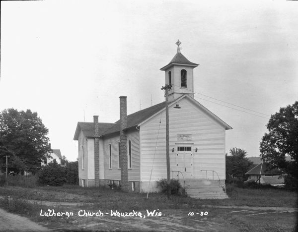View across road or path of St. Paul's Evangelical Lutheran Church, which is a wood frame building with a double door entrance with transom windows above, and arched windows and two chimneys along the left side. Above the entrance is a small, roofed belfry. The sign above the entrance reads: "St. Paul's Evangelical Lutheran Church 1873-1927."