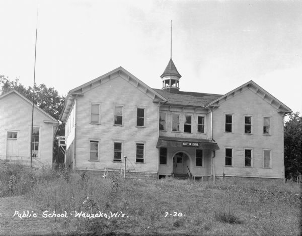 View up hill of the public school, which is a two-story wood frame building with a belfry in the center of the roof. Stairs lead to the arched entrance, and to the right of the entrance is a hand-pump. There is a merry-go-round in the schoolyard in front. A flagpole is in front of the building next door.