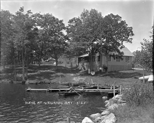 View from shoreline towards cottage at Weigands Bay, Lake Wisconsin. A pier and several small boats are along the shore, with a man posed sitting in a rowboat. Three women and a man pose under a tree near the cottage. Another cottage is in the background. Caption reads: "Scene at Weigand's Bay."