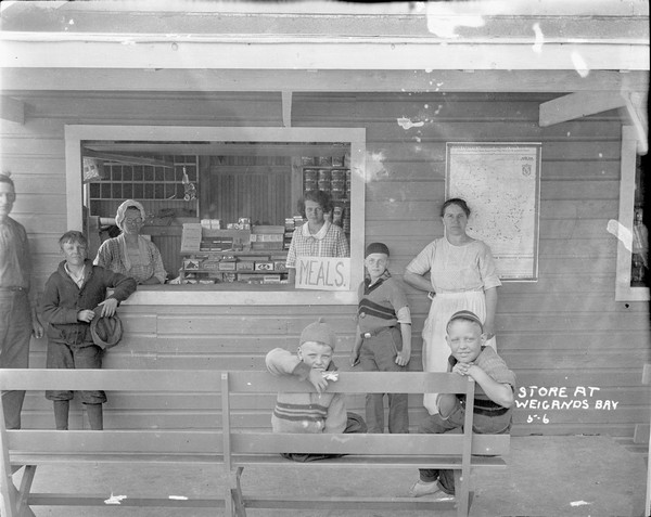 A general store counter at Weigands Bay. Two women are behind the counter, with boxes of cigars on display behind them. Other canned goods are on the shelves in the back. Standing at the open window on the left are a man and boy. On the right is woman and a boy standing in front of a Wisconsin map and near a sign on the counter that says "Meals." In the foreground sitting on a bench are two boys. The boy in the center has a bandage around his finger.