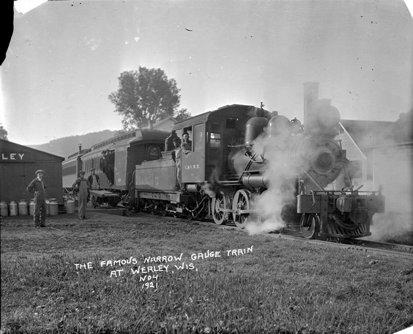 A passenger train stopped at the depot. Three men are standing on the left, one of them pushing a cart. A row of milk cans is along the side of the depot building. The engineer is leaning out the locomotive window. Two passengers stand at the open door of a train car.