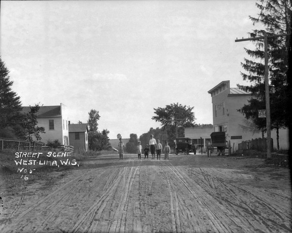 View down middle of unpaved street towards six children standing in the middle. There is a storefront on the right, with an automobile and a horse and buggy parked in front.