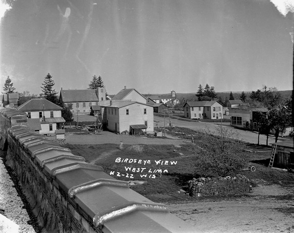 Elevated view taken from the roof of a building. Yards and dwellings in the foreground, with a church and school in the background. There are three or four children standing at the intersection. There is a large pile of cinder blocks in the foreground.